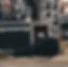 A black guitar case stands on a wooden flooring of the scene, next to boxes and music spea